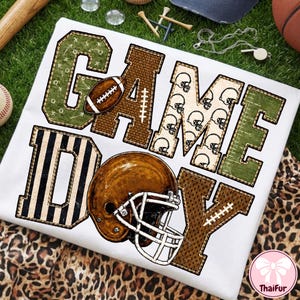 May include: A white t-shirt featuring the text GAME DAY in large block letters. The letters are filled with various football-themed patterns including green play diagrams, brown textures, and helmet icons. A brown football helmet is centered below.