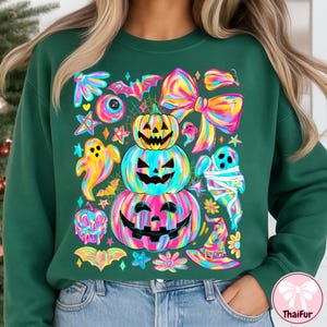 May include: A dark green sweatshirt featuring a colorful Halloween-themed design. The graphic includes stacked jack-o'-lanterns, ghosts, bats, bows, and stars in vibrant rainbow hues. The design is whimsical and festive, perfect for the spooky season.
