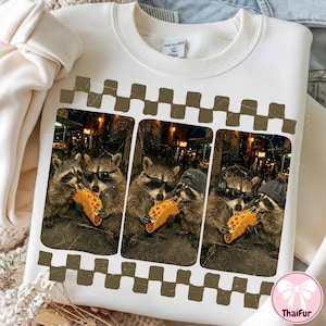 Puede incluir: Sudadera de color crema con un gráfico de tres mapaches con gafas de sol comiendo tacos. La imagen está enmarcada con un borde a cuadros marrón y blanco. Los mapaches están sobre un fondo urbano borroso.