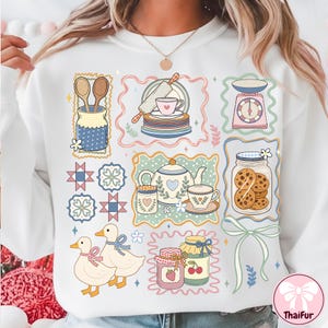 Peut inclure: Sweat-shirt blanc avec un motif color&eacute; illustrant des &eacute;l&eacute;ments de cuisine et de p&acirc;tisserie. Le motif comprend une th&eacute;i&egrave;re, des biscuits, des canards, des pots de confiture et une balance. Le motif est encadr&eacute; de bordures d&eacute;coratives.