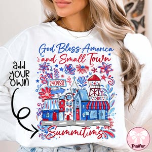 May include: White sweatshirt with a colorful graphic design. The design features the text "God Bless America and Small Town" with illustrations of buildings, flowers, and a water tower in red, white, and blue. The word "Summit, ms" is also included.