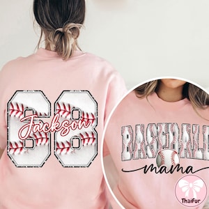 May include: A pale pink sweatshirt with baseball graphics. The left side has the number "68" with "Jackson" in script. The right side shows "Baseball Mama" with a baseball graphic. A ThaiFur logo is at the bottom.