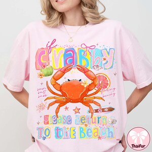 May include: Pale pink t-shirt with a vibrant graphic design. The design includes a crab, the words "If Crabby Please Return To The Beach", and beach-related illustrations. The t-shirt has a loose fit.