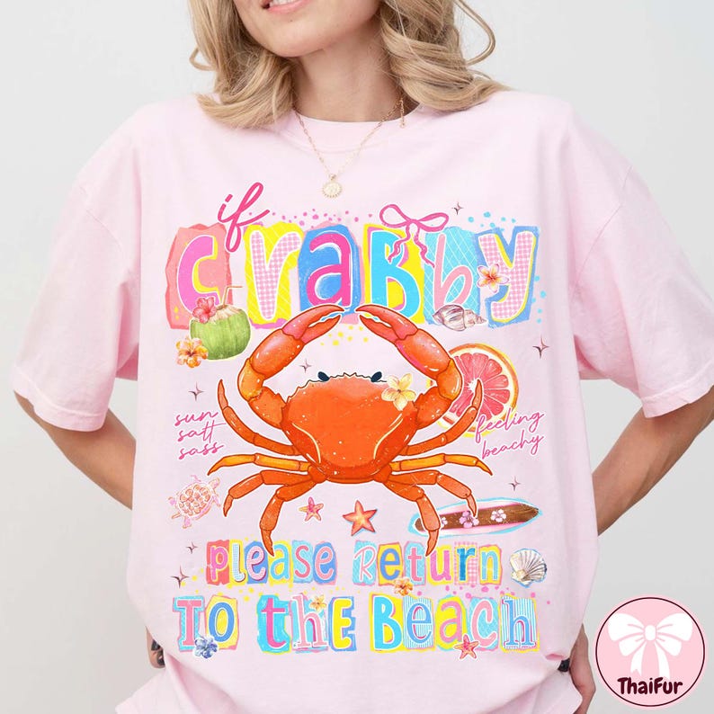 May include: Pale pink t-shirt with a vibrant graphic design. The design includes a crab, the words "If Crabby Please Return To The Beach", and beach-related illustrations. The t-shirt has a loose fit.