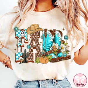May include: Cream-colored t-shirt with a 'Fall' graphic featuring a cowboy hat, turquoise boots, a pumpkin, and a cactus. The letters are brown and decorated with a checkered pattern and a cactus design. The boots are adorned with flowers.