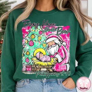 May include: Green sweatshirt with a Christmas-themed graphic. The design features Santa Claus in pink, kneeling before baby Jesus, with a decorated Christmas tree. Text reads "Every Knee Shall Bow" and "Philippians 2:10".
