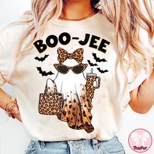 May include: Cream-colored t-shirt with a Halloween-themed graphic. The design features a ghost wearing sunglasses and a bow, carrying a leopard print bag and a cup. The text "BOO-JEE" is above the ghost, with bats in the background.
