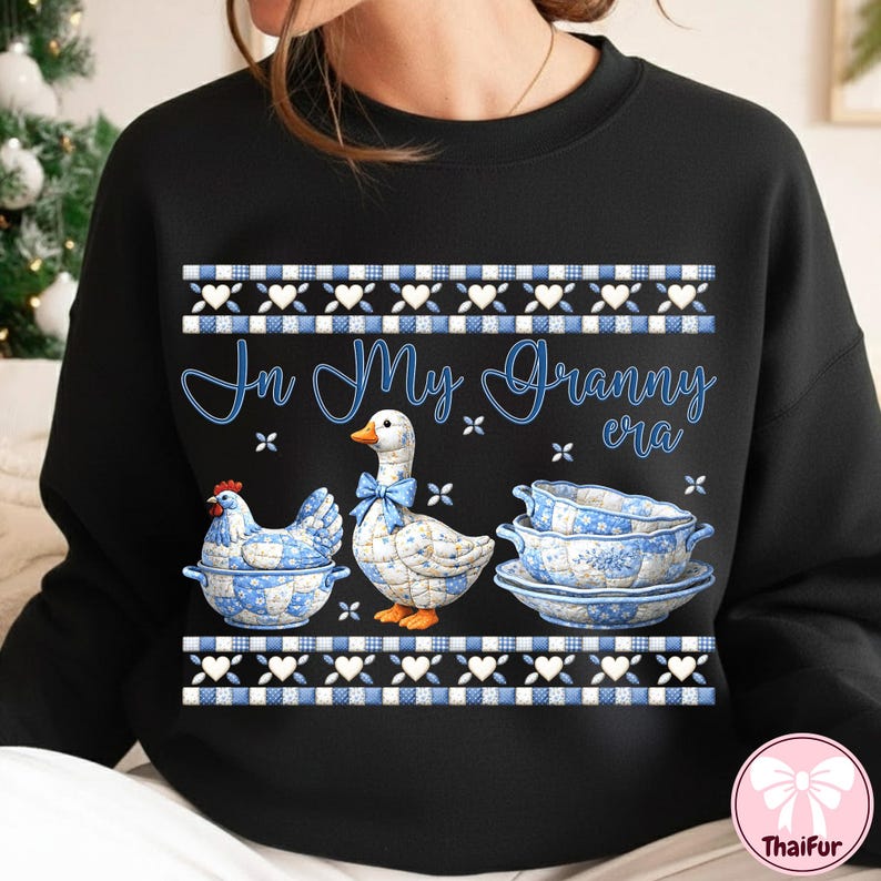 May include: Black sweatshirt with the text "In My Granny Era" in blue script. The design features a chicken, a goose with a blue bow, and a stack of blue and white floral dishes. The top and bottom of the design have a blue and white pattern.