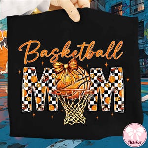 May include: Black t-shirt with the words "Basketball Mom" in orange script. The design features a basketball with a bow, a net, and the letters "MOM" with a checkered pattern and basketball details.