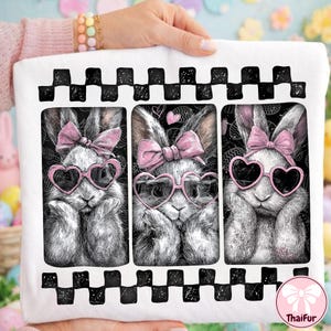 May include: White square pillow with three bunny portraits. Each bunny wears pink heart-shaped sunglasses and a pink bow. The top and bottom edges have a black and white checkered pattern. The pillow is held by a person.