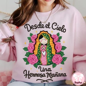 May include: A light pink sweatshirt featuring a graphic of a religious figure surrounded by pink roses and green leaves. The text "Desde el Cielo Una Hermosa Mañana" is written in black script above and below the graphic.