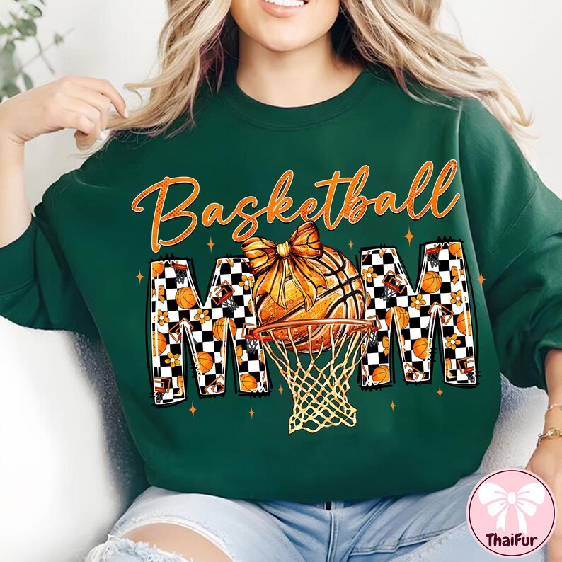 May include: Dark green sweatshirt with the words "Basketball Mom" in orange script and block letters. The "MOM" letters have a checkered pattern with basketballs and flowers. A basketball with a bow sits above a basketball hoop.