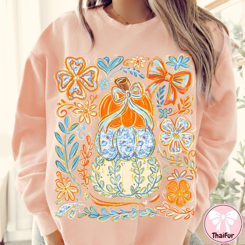 May include: A light pink sweatshirt featuring a stacked pumpkin design with floral and bow accents. The pumpkins are orange, blue, and yellow, with blue and orange floral and leaf details. The design is surrounded by orange and blue floral elements.