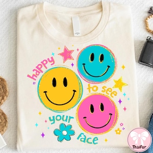 Op de afbeelding: Crèmekleurig T-shirt met een grafisch ontwerp met drie smiley-gezichten in geel, blauw en roze. De tekst "happy to see your face" staat rond de gezichten, samen met sterren en een bloem.