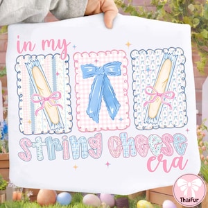May include: White fabric with the text "in my String Cheese era" in a whimsical font. The design includes three framed illustrations: two string cheese sticks tied with pink bows and a blue bow. Pastel colors.