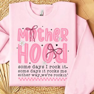 May include: Pink sweatshirt with the text "Mother Hood" in pink letters with a pink bow and flowers. The text below reads "some days I rock it, some days it rocks me, either way, we're rockin'."