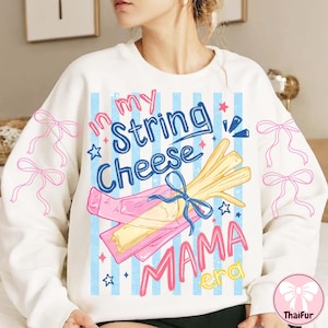 May include: White sweatshirt with a graphic design featuring the text "in my String Cheese Mama era". The design includes a cartoon illustration of string cheese, stars, and pink and blue accents. Pink bows are printed on the sleeves.