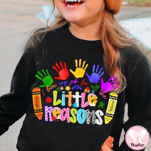 May include: Black sweatshirt with a colorful graphic design. The design includes handprints in green, red, yellow, blue, and purple, along with the text "I love my job for all the Little Reasons" and crayon illustrations.