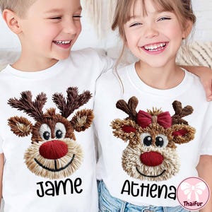 Op de afbeelding: Twee kinderen dragen witte T-shirts met geborduurde rendiergezichten. De rendieren hebben bruine geweien, rode neuzen en lachende monden. Eén shirt zegt "Jame" en de andere "Athena" in zwarte letters.