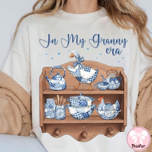 K&ouml;nnte beinhalten: Cremefarbenes T-Shirt mit dem Text "In My Granny era" in blauer Schrift. Das Shirt zeigt ein Holzregal-Design mit blau-wei&szlig; kariertem K&uuml;chengeschirr und einer Gans.