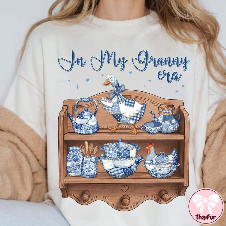 K&ouml;nnte beinhalten: Cremefarbenes T-Shirt mit dem Text "In My Granny era" in blauer Schrift. Das Shirt zeigt ein Holzregal-Design mit blau-wei&szlig; kariertem K&uuml;chengeschirr und einer Gans.