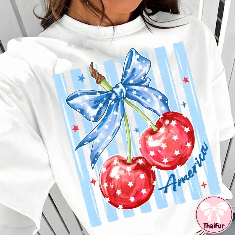 May include: White t-shirt featuring a patriotic design with two red cherries adorned with white stars. A blue bow with white stars and blue and white striped background. The word "America" is written in blue.