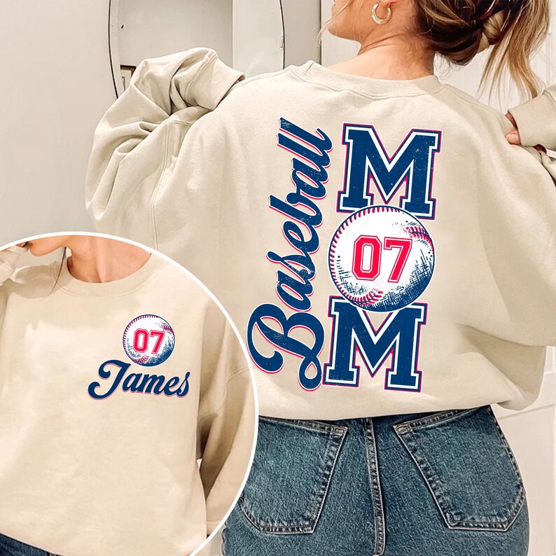 May include: Beige sweatshirt with a baseball-themed design on the back. The design features a baseball with the number 07, the word "Baseball" and the word "MOM" in blue and red lettering. The front of the sweatshirt has a smaller version of the baseball design with the name "James" underneath.
