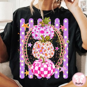 May include: A black t-shirt featuring a graphic of three stacked pumpkins inside a decorative oval frame. The top pumpkin is purple with spots, the middle has an orange leopard print, and the bottom has a pink checkerboard pattern with stars.