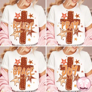May include: A white t-shirt featuring a brown polka-dot cross graphic surrounded by orange and red stars. The design includes the words Believe, Pray, Grace, or Joy, paired with Bible verse references like John 14:1, Colossians 4:2, 2 Cor 12:9, or John 16:22.