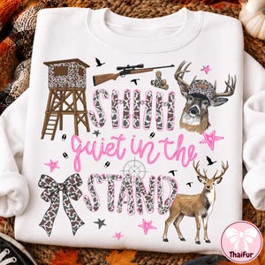 Könnte beinhalten: Ein weißes Sweatshirt mit einem Grafikdruck, der einen Hochsitz, ein Gewehr, zwei Hirsche, eine Schleife und den Text SHHH quiet in the STAND zeigt. Das Design umfasst Tarnmuster, rosa Sterne und Vogel-Silhouetten auf der Vorderseite.