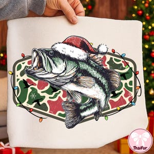 Op de afbeelding: Een decoratief kussen met een gedetailleerde illustratie van een largemouth bass met een kerstmuts. De vis staat tegen een camouflagepatroon met zwarte vogels, omringd door een slinger van kleurrijke kerstverlichting. Het kussen wordt door een persoon vastgehouden.