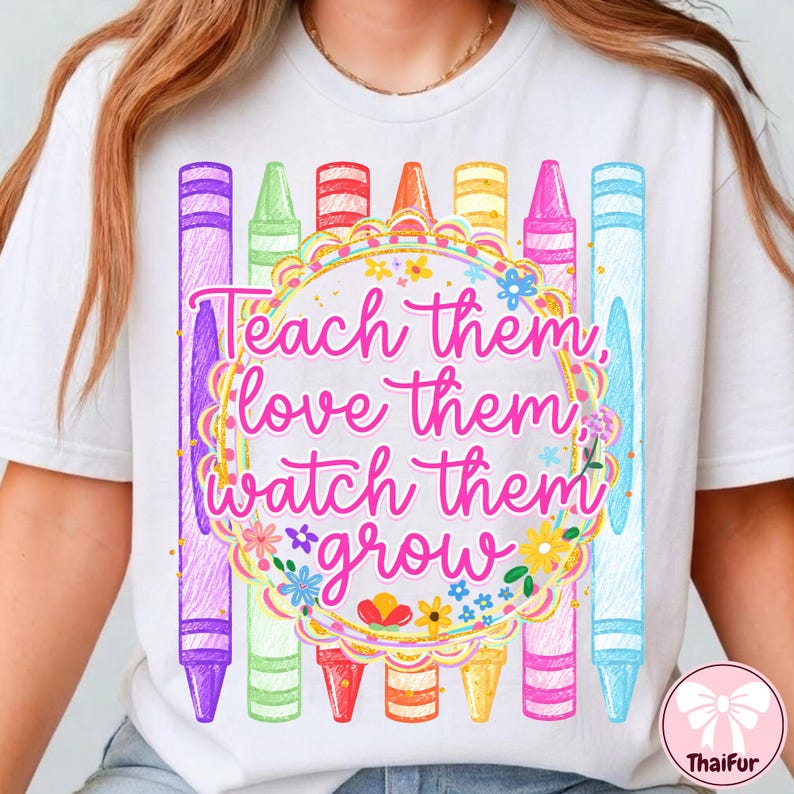 May include: A white t-shirt featuring a graphic of colorful crayons arranged vertically behind a circular frame. The text "Teach them, love them, watch them grow" is printed in pink cursive script inside the frame, surrounded by small floral accents.