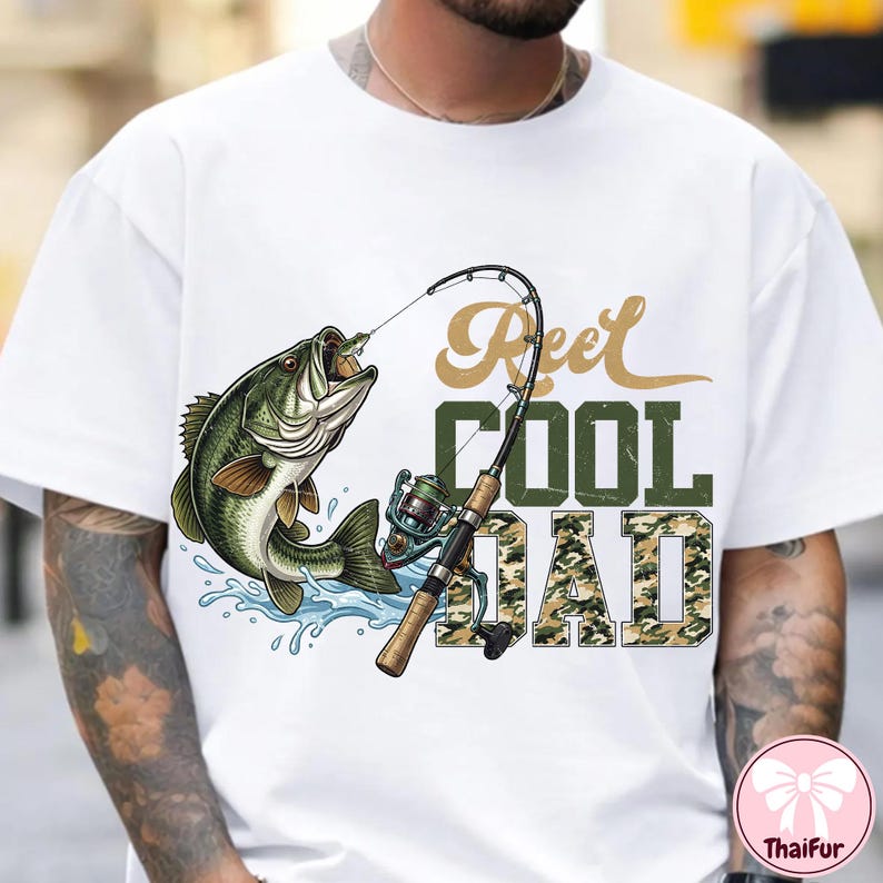 May include: White t-shirt featuring a graphic of a largemouth bass caught on a fishing rod. The text "Reel Cool Dad" is printed in a combination of gold and camouflage green.