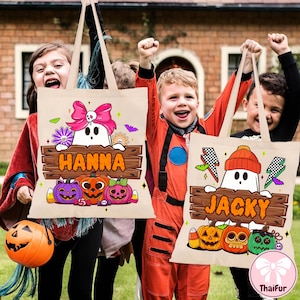 May include: Beige Halloween tote bags with festive designs. One features a ghost with a pink bow, the name "HANNA", and pumpkins. The other has a ghost wearing a beanie, the name "JACKY", and pumpkins. Both have candy corn accents.