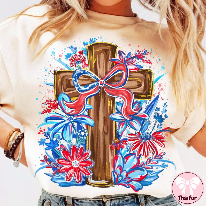 May include: Cream-colored t-shirt featuring a brown cross adorned with a red, white, and blue bow. The cross is surrounded by red, white, and blue floral designs, creating a patriotic and decorative theme. The shirt has a round neckline.