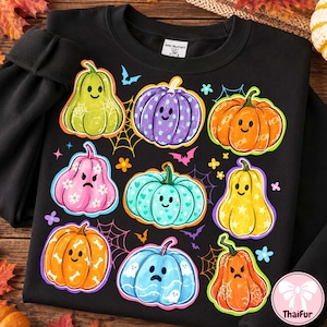 May include: A black crewneck sweatshirt featuring a colorful graphic print of nine stylized pumpkins with various facial expressions, patterns, and colors, interspersed with small illustrations of spiderwebs, bats, and flowers.