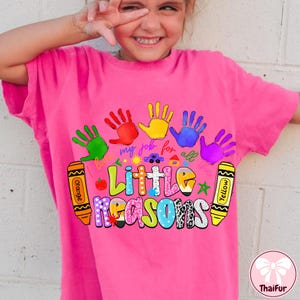 May include: A pink t-shirt featuring a colorful design with handprints, crayons, and the text "Little Reasons." The design includes the phrase "my job for all the." The shirt is a vibrant and playful design.