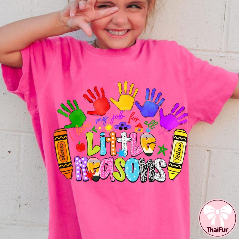 May include: A pink t-shirt featuring a colorful design with handprints, crayons, and the text "Little Reasons." The design includes the phrase "my job for all the." The shirt is a vibrant and playful design.