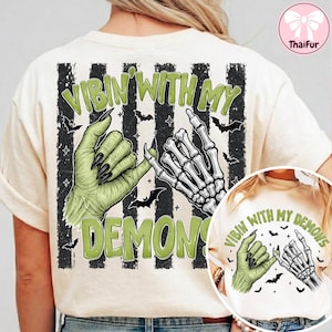 May include: A cream-colored t-shirt featuring a graphic of a green monster hand and a white skeleton hand performing a pinky promise. The design includes black vertical stripes, small bats, and the text VIBIN WITH MY DEMONS in green.