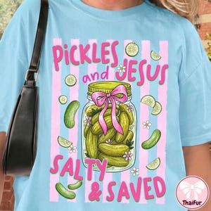 Könnte beinhalten: Hellblaues T-Shirt mit dem Text "Pickles and Jesus Salty & Saved". Die Grafik zeigt ein Glas mit Essiggurken mit einer rosa Schleife, umgeben von Gurkenscheiben und Blumen, vor rosa vertikalen Streifen.