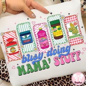 May include: White pillow with colorful graphics and text. The design includes images of products and the words "busy doing MAMA' STUFF". The graphics include a bottle, jar, and package with text in Spanish.