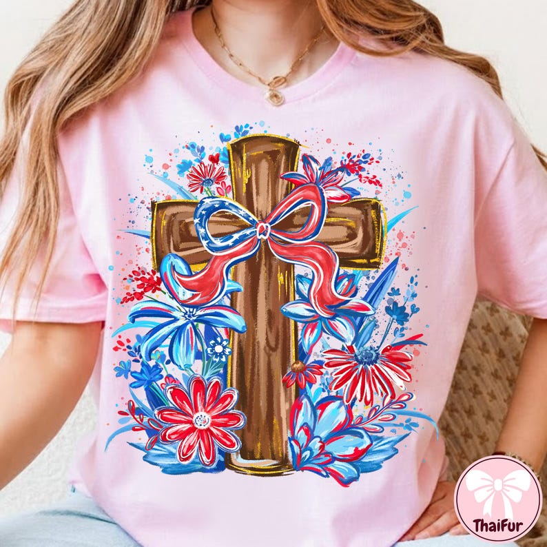 May include: A light pink t-shirt featuring a brown cross adorned with a red, white, and blue bow. The cross is surrounded by red and blue floral designs, creating a patriotic and decorative theme.