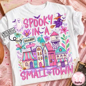 Op de afbeelding: Een wit t-shirt met een kleurrijke afbeelding met de tekst SPOOKY IN A SMALL TOWN en een bordje met 666889. Het ontwerp bevat een heks op een bezemsteel, geesten, vleermuizen, spinnenwebben, bloemen en een rij gebouwen in roze en paarse tinten.