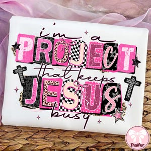 May include: White t-shirt with a graphic design in pink, black, and white. The design includes the text "I'm a PROJECT that keeps JESUS busy" in a block letter style, with stars, bows, and cross accents.