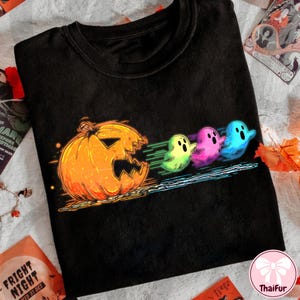 Pode incluir: Camiseta preta com um design de Halloween. Uma abóbora laranja é retratada com uma fileira de fantasmas coloridos atrás dela. Os fantasmas são em tons de verde, rosa e azul. O design tem um efeito de brilho neon.