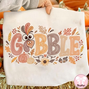 May include: A white t-shirt featuring the word GOBBLE in large, multi-colored, line-art style letters. The letter O is replaced by a turkey face. The design includes orange, brown, and yellow pumpkins, leaves, acorns, and a sunflower.