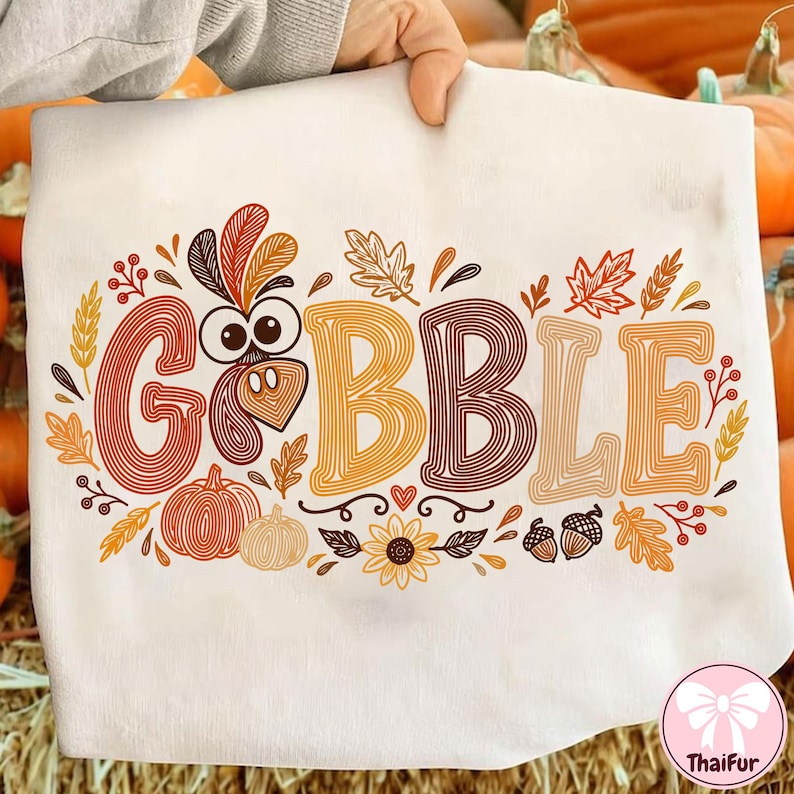 May include: A white t-shirt featuring the word GOBBLE in large, multi-colored, line-art style letters. The letter O is replaced by a turkey face. The design includes orange, brown, and yellow pumpkins, leaves, acorns, and a sunflower.