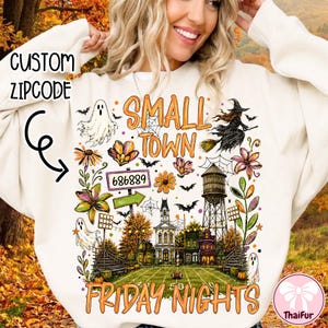 May include: A cream-colored sweatshirt featuring a graphic print with the text "SMALL TOWN FRIDAY NIGHTS" and "686889". The design includes a football field, a water tower, a witch on a broomstick, ghosts, bats, spiderwebs, and colorful flowers.