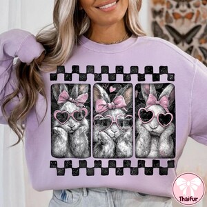 May include: A light purple sweatshirt featuring a graphic of three bunnies wearing heart-shaped sunglasses and pink bows. The bunnies are in a black and white checkered frame. The ThaiFur logo is in the bottom right corner.