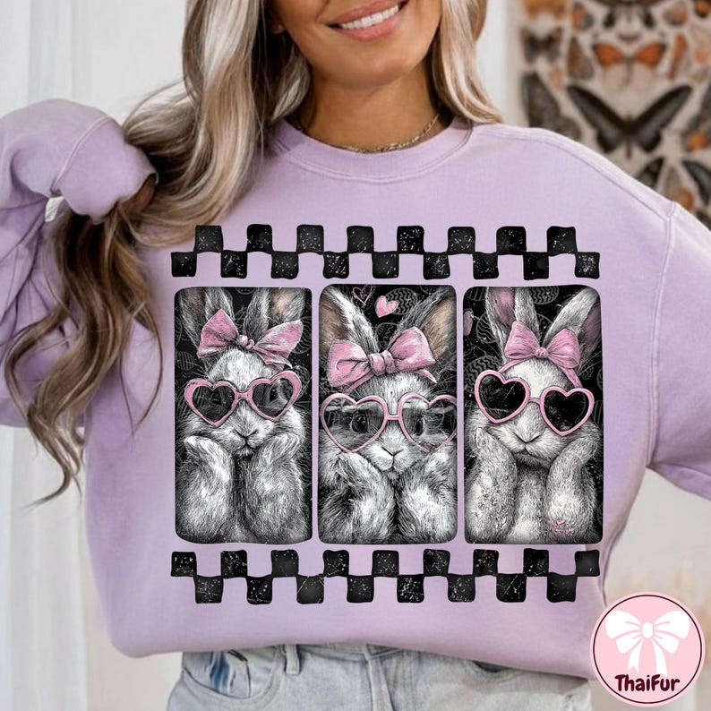 May include: A light purple sweatshirt featuring a graphic of three bunnies wearing heart-shaped sunglasses and pink bows. The bunnies are in a black and white checkered frame. The ThaiFur logo is in the bottom right corner.